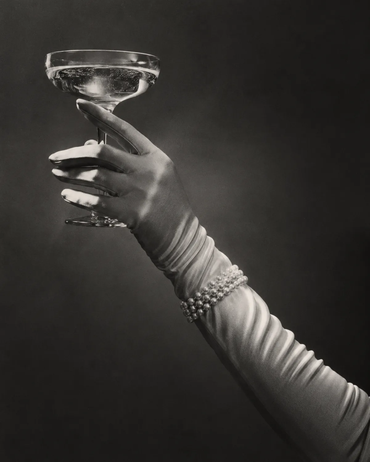 Schwarzweiß-Fotografie: behandschuhte Hand mit Perlenarmband hält eine Coupe